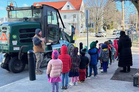 classroom of early education students stand in front of the Capuano with their teacher, listening to a DPW worker talk about the street cleaner he operates