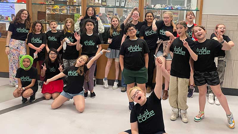 Girls Who Code participants in matching program t-shirts and teacher pose in front of Fablab