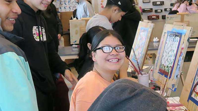 Photo of smiling student in front of her easel with other students looking at her artwork and smiling
