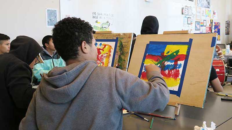 Photo of Student artists painting at their easels as they listen to a student drumming performance