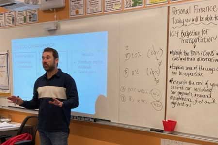 Photo of guest speaker in Somerville High School personal finance 1 class. He is in jeans and a sweater, gesticulating in front of a white board.