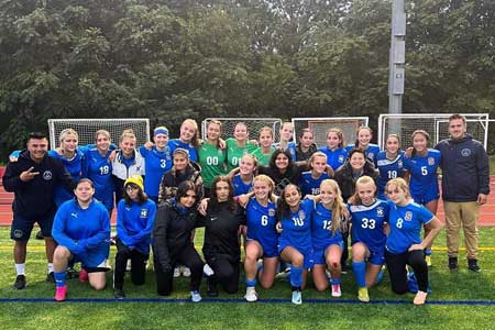 Girls Varsity Soccer Team photo with coaches on a soccer pitch against a leafy, green backdrop
