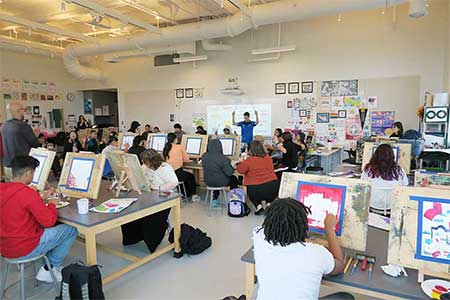 Photo in the art studio of students painting while a drumming class performs. Staff and other students stand around the edge of the photo.