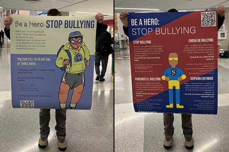 Two photos of the same student holding up each poster design. He is wearing hiking boots and standing in the atrium near the entrance to the high school.