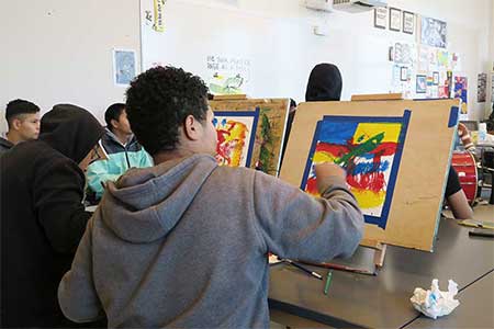Students sit in front of easels painting with student drummers in the background. The students are in an art studio in the high school.