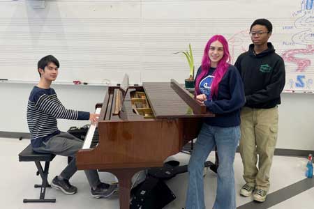 Three student musicians around a piano in the orchestra room at the high school