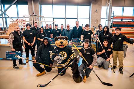 Photo of carpentry students, representatives from the Bruins Foundation, and Bosch Tools representatives posing with donated hockey sticks and power tools in the carpentry studio