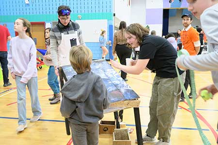 Photo of students in the Kennedy gym with two middle grades students showing a younger student how to use a game they designed and built. There are several people and students moving around in the background.