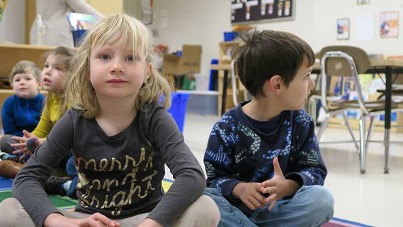 Two kindergarten students sit in front of other students, criss cross apple sauce on a rug in the K classroom.