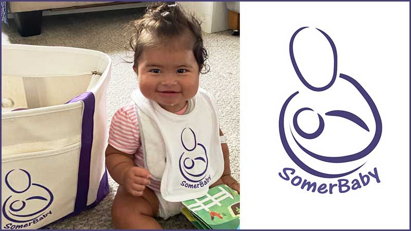 Baby holding a soft book, next to a branded tote on a carpet. Somerbaby logo