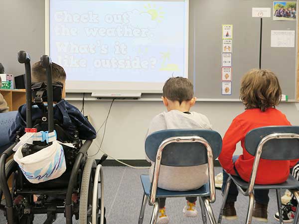 Photo of three students sitting and looking at a smartboard with a question about the weather. The student on the left is in a wheelchair. 