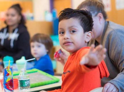 Student, reaches an arm to photographer. Sits in foreground in an orange shirt with two adults and a child behind him in a PK classroom