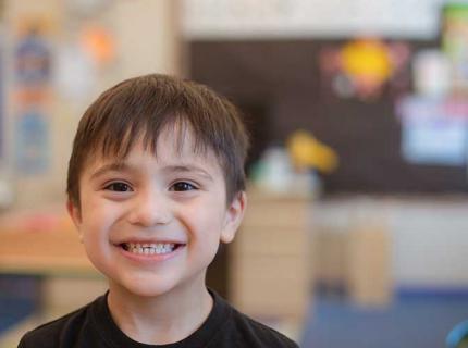 Small boy in a black t-shirt smiling at the camera in a classroom.